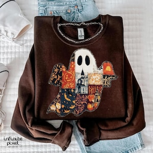 Puede incluir: Una sudadera marr&oacute;n con un aplique de fantasma hecho de varias telas estampadas. El fantasma tiene una cara blanca con ojos negros y un cuerpo de patchwork con dise&ntilde;os de Halloween. Una cadena de plata descansa sobre la sudadera.