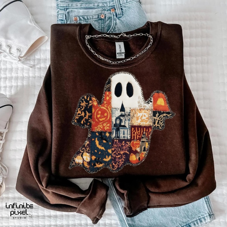 Puede incluir: Una sudadera marr&oacute;n con un aplique de fantasma hecho de varias telas estampadas. El fantasma tiene una cara blanca con ojos negros y un cuerpo de patchwork con dise&ntilde;os de Halloween. Una cadena de plata descansa sobre la sudadera.