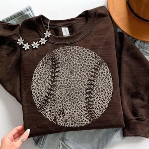 May include: A brown sweatshirt featuring a baseball design with a leopard print pattern. The baseball has white stitching details. A silver flower necklace is draped on the sweatshirt. The sweatshirt is on top of a pair of jeans.
