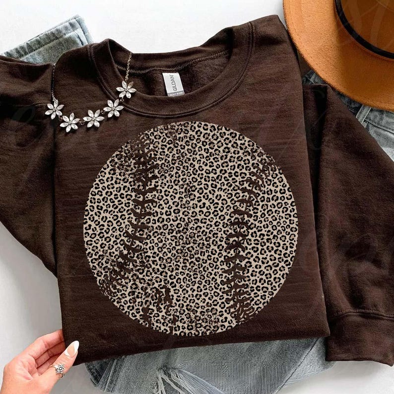 May include: A brown sweatshirt featuring a baseball design with a leopard print pattern. The baseball has white stitching details. A silver flower necklace is draped on the sweatshirt. The sweatshirt is on top of a pair of jeans.