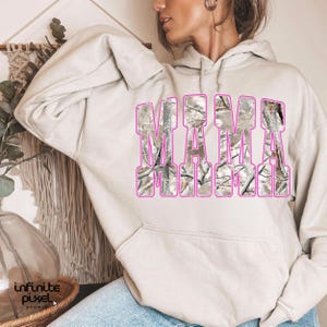 May include: A beige hoodie with the word "MAMA" in large, block letters across the front. The letters are filled with a camouflage pattern and outlined in pink. The hoodie has a drawstring hood and long sleeves.