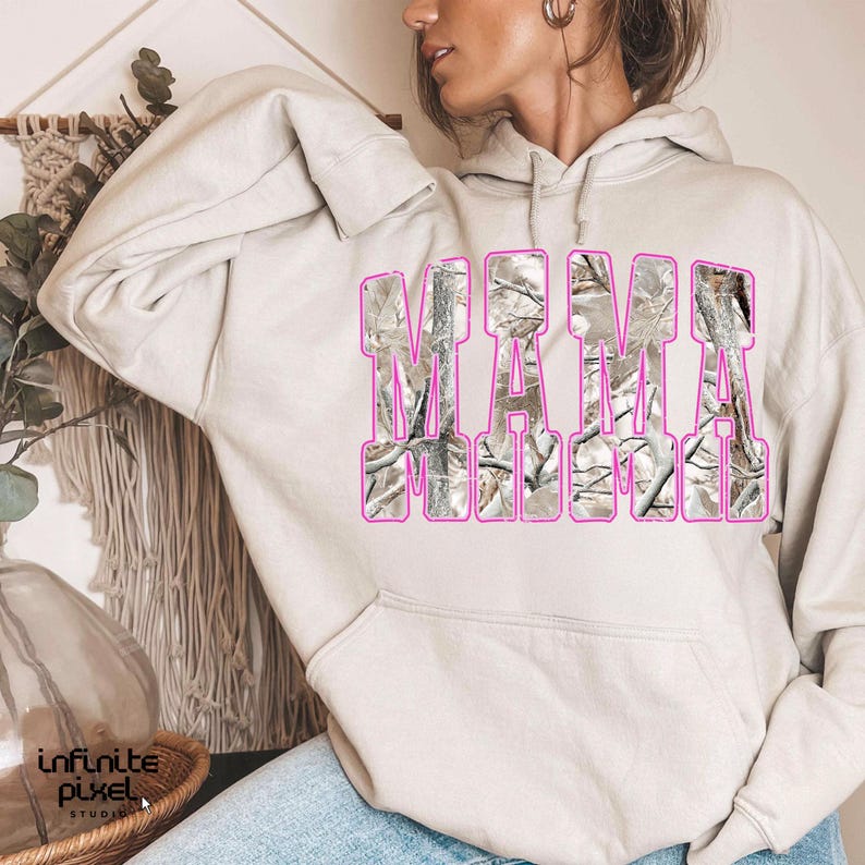 May include: A beige hoodie with the word "MAMA" in large, block letters across the front. The letters are filled with a camouflage pattern and outlined in pink. The hoodie has a drawstring hood and long sleeves.