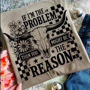 May include: A tan t-shirt with a black graphic of a bull skull, flowers, and the text "IF I'M THE PROBLEM THEN YOU MIGHT BE THE REASON." The design includes a checkered pattern and stars.