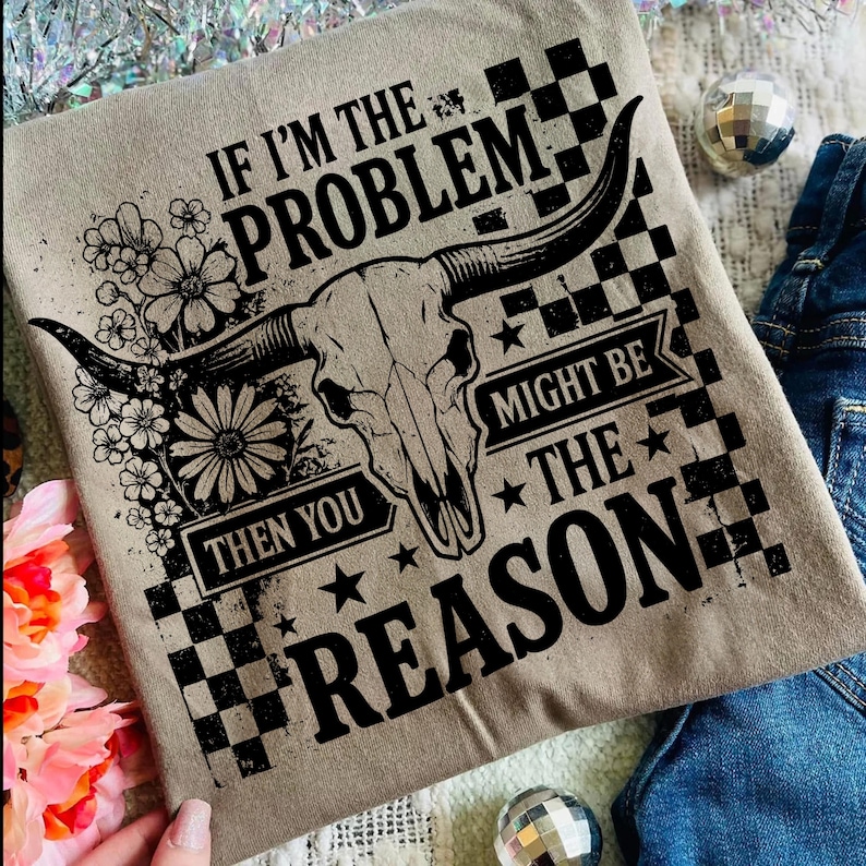May include: A tan t-shirt with a black graphic of a bull skull, flowers, and the text "IF I'M THE PROBLEM THEN YOU MIGHT BE THE REASON." The design includes a checkered pattern and stars.