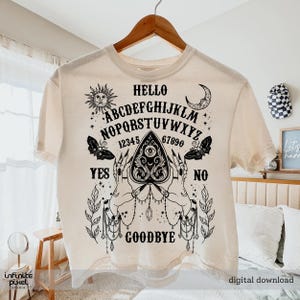 May include: A cream-coloured t-shirt featuring a black Ouija board design. The design includes the alphabet, numbers, and the words "Hello", "Yes", "No", and "Goodbye". The graphic also features a sun, moon, butterflies, and hands.