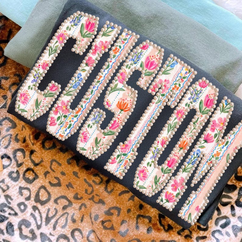 Puede incluir: Una camiseta gris oscuro con la palabra "Gigi" en un dise&ntilde;o floral. Las letras est&aacute;n delineadas con perlas y rellenas de flores rosas, naranjas y azules. La camiseta est&aacute; doblada sobre una superficie con estampado de leopardo.