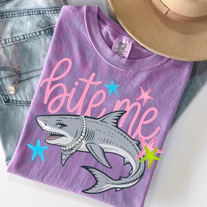 May include: A purple Comfort Colors t-shirt with the phrase "bite me" in pink script, featuring a cartoon shark wearing a pearl necklace. The shark is gray with white teeth and is surrounded by blue and yellow stars.
