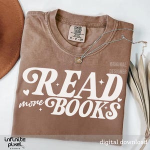 Peut inclure: Un t-shirt marron avec les mots "READ more BOOKS" en lettres blanches stylisées. Le t-shirt a un col rond et des manches courtes. Un collier argenté avec un pendentif est drapé sur le t-shirt. Les mots "ORIGINAL DESIGNER" sont imprimés sur le t-shirt.