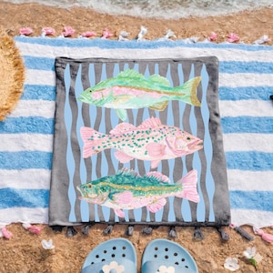 May include: A beach towel with three colorful fish illustrations. The fish are in shades of green, pink, and gold, set against a light blue wavy background. The towel is gray with a blue and white striped border.