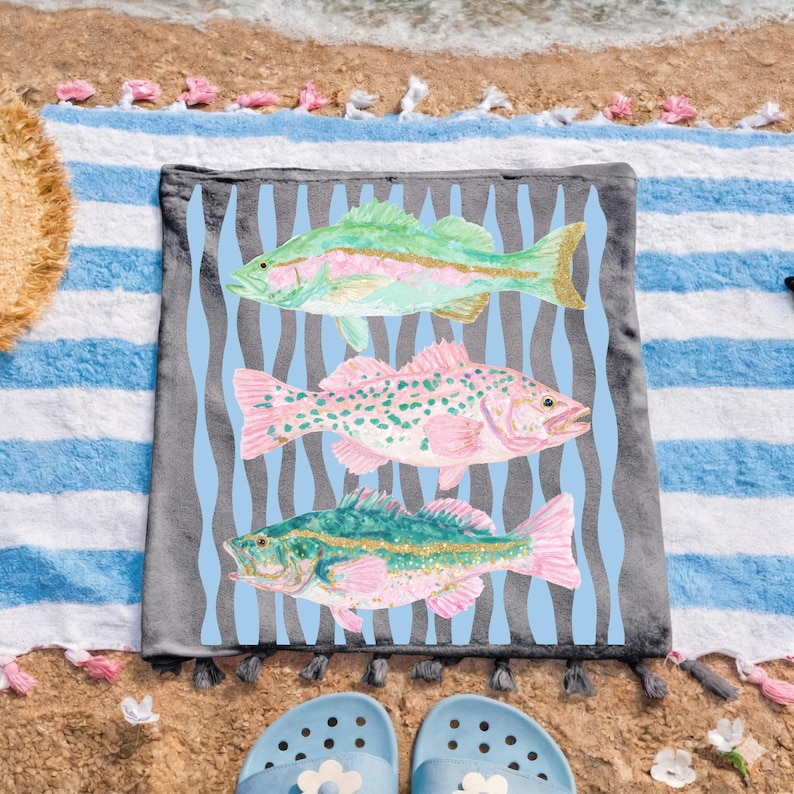 May include: A beach towel with three colorful fish illustrations. The fish are in shades of green, pink, and gold, set against a light blue wavy background. The towel is gray with a blue and white striped border.