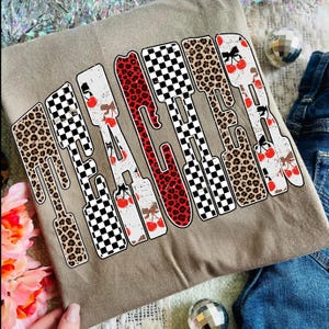 May include: A tan t-shirt with the word "TEACHER" in a bold, stylized font. The letters feature a mix of patterns, including leopard print, red, and black and white checkerboard, with cherry accents. The shirt is paired with denim.