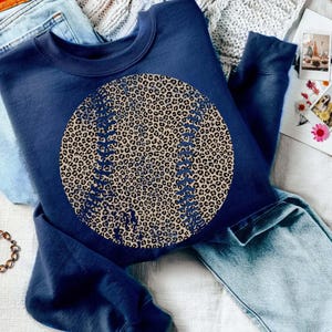 May include: Navy blue sweatshirt featuring a large baseball design with a leopard print pattern. The baseball has detailed stitching. The sweatshirt is paired with light blue jeans and a gold bracelet. A notebook and photos are in the background.