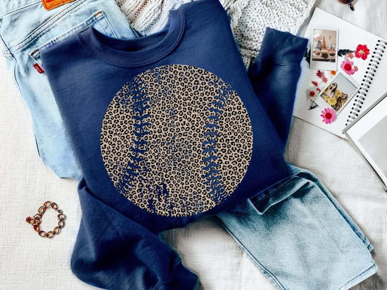 May include: Navy blue sweatshirt featuring a large baseball design with a leopard print pattern. The baseball has detailed stitching. The sweatshirt is paired with light blue jeans and a gold bracelet. A notebook and photos are in the background.