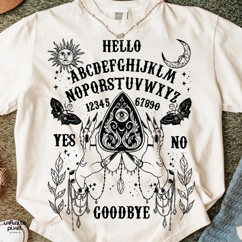May include: Cream-coloured t-shirt with a black Ouija board design. The graphic includes the alphabet, numbers, sun, moon, butterflies, and the words "Hello", "Yes", "No", and "Goodbye". The design is centered on the front of the shirt.
