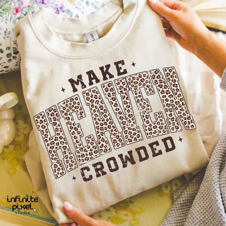 May include: A beige sweatshirt with the text "MAKE HEAVEN CROWDED" in brown lettering. The word "HEAVEN" is filled with a leopard print pattern. The sweatshirt is folded and held by a person.
