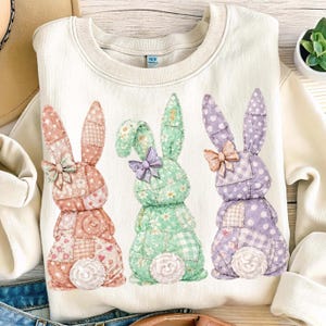 May include: Cream-colored sweatshirt featuring three patchwork bunny designs in pink, green, and purple. Each bunny has a bow and a fluffy tail. The bunnies are made of various patterned fabrics, including polka dots and gingham.