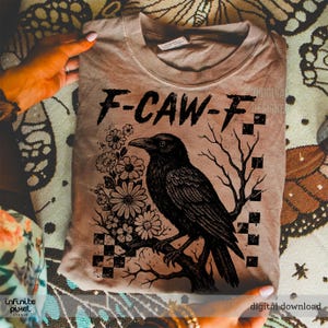 Puede incluir: Camiseta color beige con un dise&ntilde;o de cuervo negro posado en una rama, rodeado de flores y un gr&aacute;fico de &aacute;rbol. El texto "F-CAW-F" est&aacute; encima del cuervo. La camiseta est&aacute; doblada y sostenida por manos.