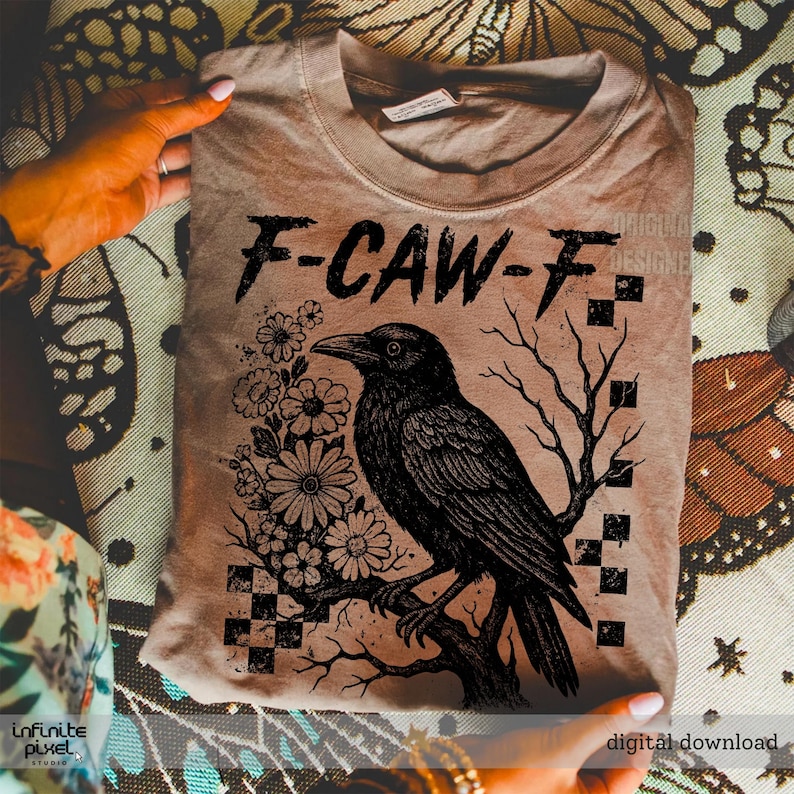 Puede incluir: Camiseta color beige con un dise&ntilde;o de cuervo negro posado en una rama, rodeado de flores y un gr&aacute;fico de &aacute;rbol. El texto "F-CAW-F" est&aacute; encima del cuervo. La camiseta est&aacute; doblada y sostenida por manos.