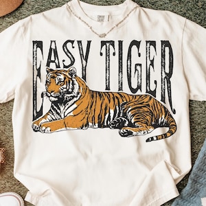 May include: Cream-coloured t-shirt with a detailed illustration of a tiger with orange and black stripes. The text "EASY TIGER" is printed above the tiger in a bold, black font. The shirt is a Comfort Colors brand.
