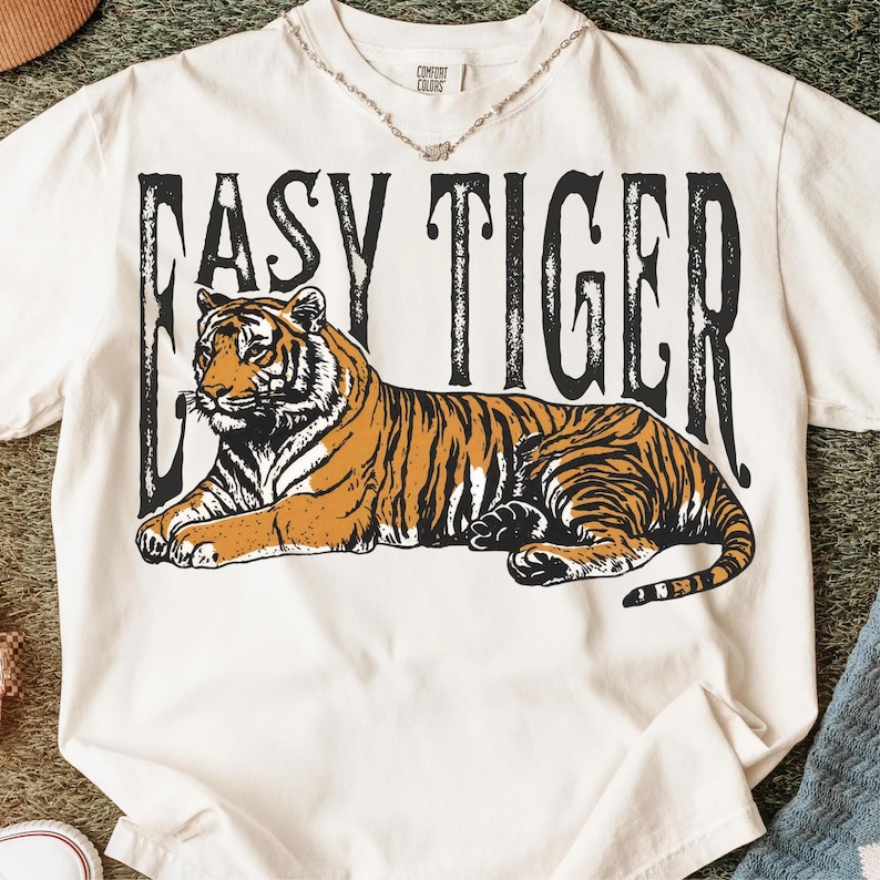 May include: Cream-coloured t-shirt with a detailed illustration of a tiger with orange and black stripes. The text "EASY TIGER" is printed above the tiger in a bold, black font. The shirt is a Comfort Colors brand.