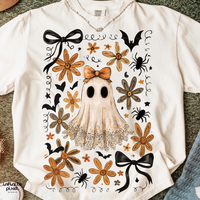 May include: A cream-coloured t-shirt with a Halloween-themed design. The graphic features a ghost with a bow, flowers, bats, spiders, and bows. The shirt has a Comfort Colors label.