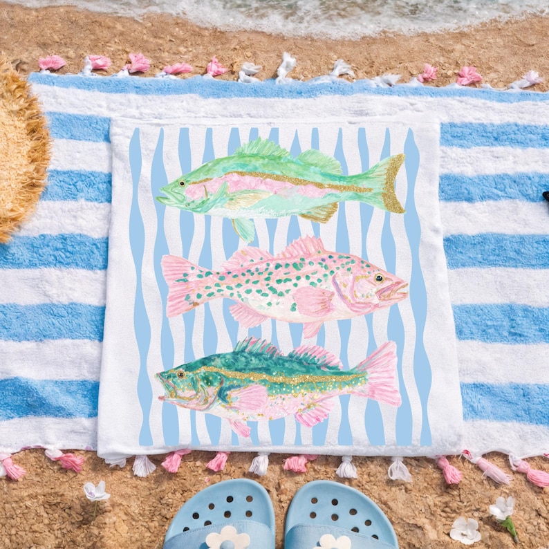 May include: A white beach towel with a blue and white striped border and a central design of three colorful fish. The fish are green, pink, and blue, with gold accents. The towel is on a sandy beach with blue sandals.