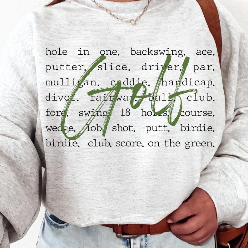May include: A gray sweatshirt with the word "golf" written in green cursive on the front. The word is surrounded by a list of golf terms, including "hole in one", "backswing", "ace", "putter", "slice", "driver", "par", "mulligan", "caddie", "handicap", "divot", "fairway", "ball", "club", "fore", "swing", "18 holes", "course", "wedge", "lob shot", "putt", "birdie", "birdie", "club", "score", and "on the green".