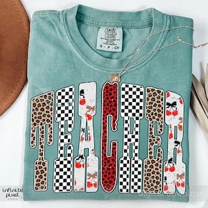 May include: A teal Comfort Colors t-shirt with the word "TEACHER" in a bold, stylized font. The letters feature a mix of leopard print, checkered patterns, and cherry designs. A gold necklace with a pendant rests on the shirt.
