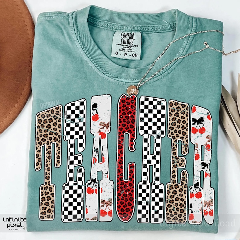 May include: A teal Comfort Colors t-shirt with the word "TEACHER" in a bold, stylized font. The letters feature a mix of leopard print, checkered patterns, and cherry designs. A gold necklace with a pendant rests on the shirt.