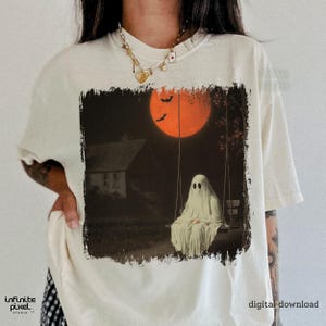May include: Cream-colored t-shirt featuring a spooky Halloween graphic. The design depicts a ghost on a swing set under an orange moon, with bats and a house in the background. The words "digital download" are printed on the shirt.