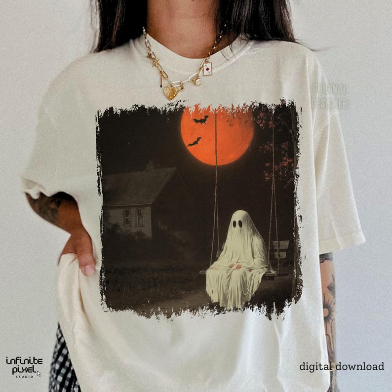 May include: Cream-colored t-shirt featuring a spooky Halloween graphic. The design depicts a ghost on a swing set under an orange moon, with bats and a house in the background. The words "digital download" are printed on the shirt.