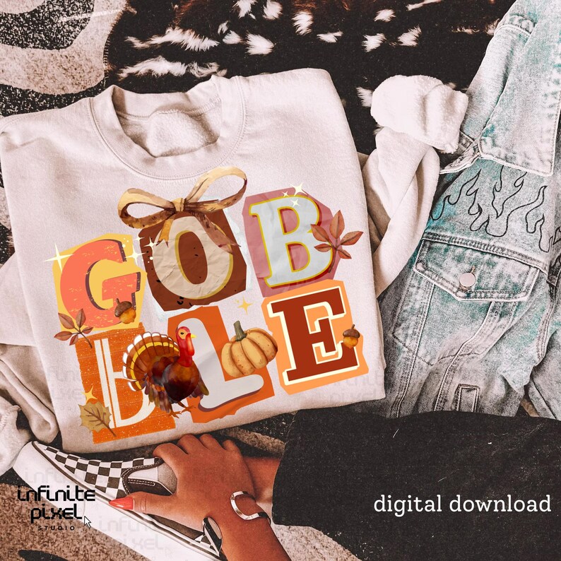 May include: Cream-colored sweatshirt with the word "GOBBLE" in colorful, fall-themed lettering. The design includes a turkey, pumpkin, and autumn leaves. A denim jacket and checkered sneakers are visible.