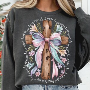 May include: A gray sweatshirt with a graphic of a wooden cross with a pink and blue bow and floral accents. The text "He is risen. He is risen. He is risen. He is risen. He is risen. He is risen. He is risen. He is risen. He is risen." is printed around the cross.