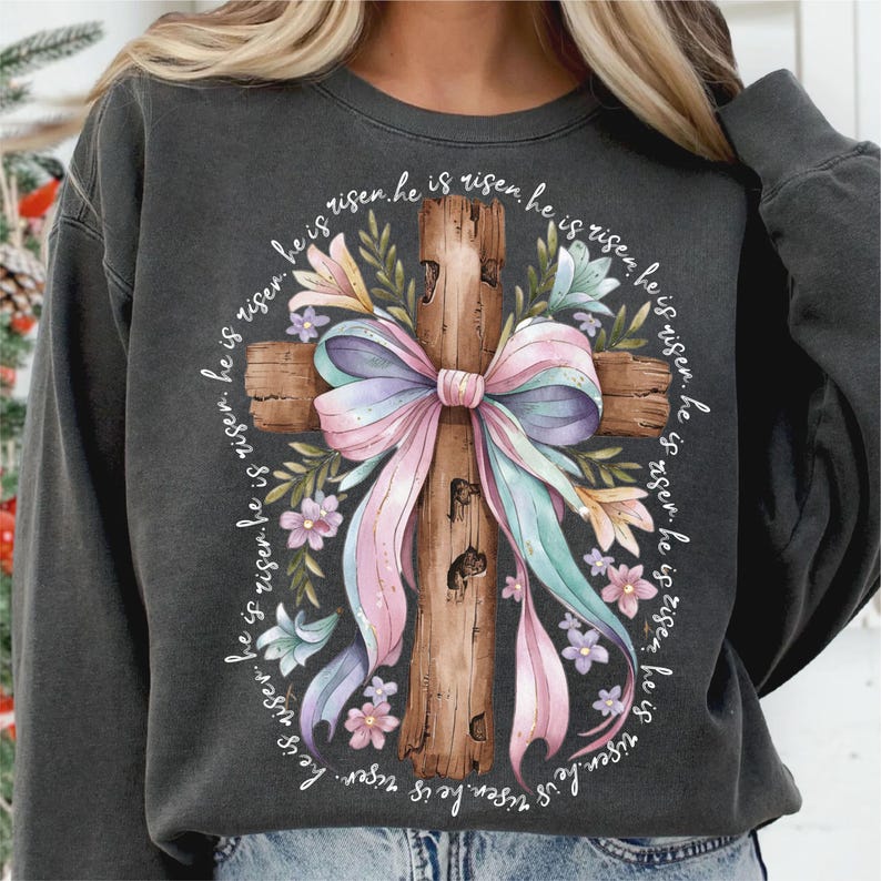 May include: A gray sweatshirt with a graphic of a wooden cross with a pink and blue bow and floral accents. The text "He is risen. He is risen. He is risen. He is risen. He is risen. He is risen. He is risen. He is risen. He is risen." is printed around the cross.