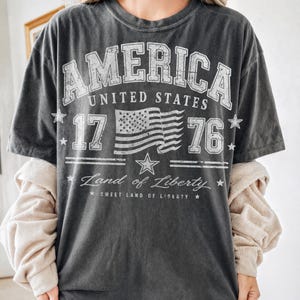 May include: Dark gray t-shirt with a distressed white graphic. The graphic includes the word "AMERICA", "UNITED STATES", the number "1776", a waving American flag, and the phrase "Land of Liberty".