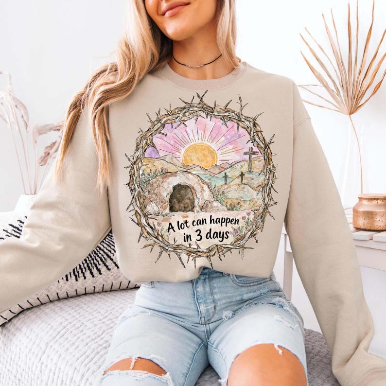 May include: Beige sweatshirt featuring a graphic of a sunrise over a tomb, with the text "A lot can happen in 3 days." The design is framed by a thorny wreath. The sweatshirt is a neutral color.