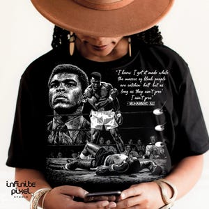 May include: Black t-shirt featuring a black and white graphic of Muhammad Ali in the boxing ring. The design includes a portrait of Ali and a quote: "I know I got it made while the masses of black people are catchin' hell but as long as they ain't free I ain't free."