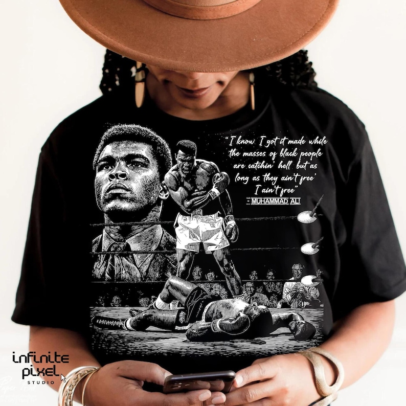 May include: Black t-shirt featuring a black and white graphic of Muhammad Ali in the boxing ring. The design includes a portrait of Ali and a quote: "I know I got it made while the masses of black people are catchin' hell but as long as they ain't free I ain't free."