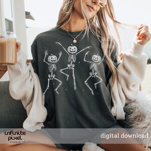 May include: Dark gray sweatshirt featuring a white graphic of three dancing skeletons with pumpkin heads. The phrase "digital download" is visible in the lower right corner. The sweatshirt is part of a Halloween-themed collection.