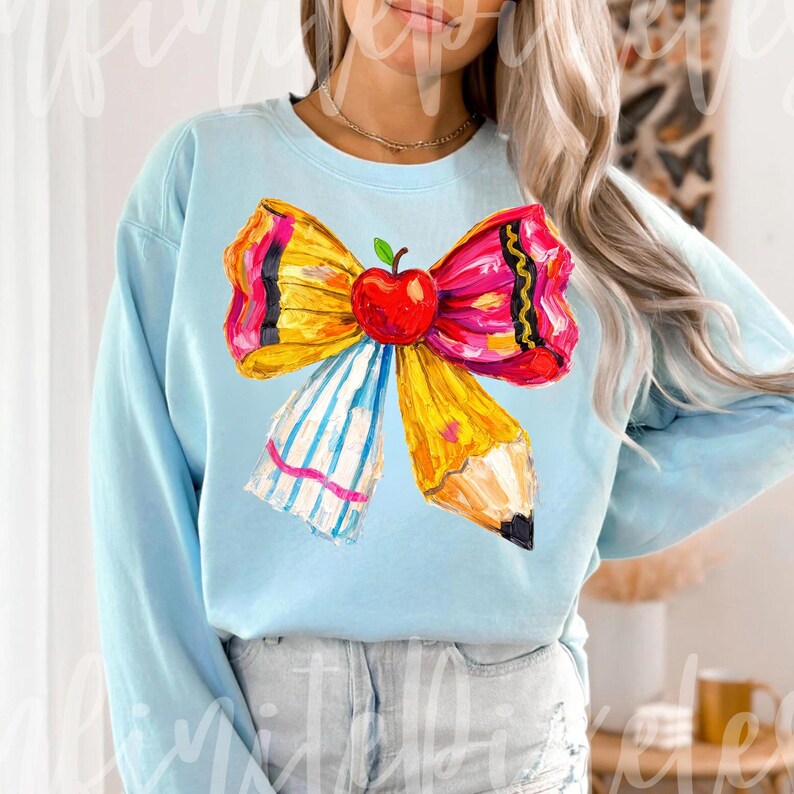 May include: Light blue sweatshirt featuring a colorful bow design with a red apple, yellow pencil, and striped ribbon. The bow is a vibrant mix of yellow, pink, and black, creating a playful and eye-catching graphic. The sweatshirt is a comfortable, casual style.
