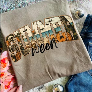 May include: A light brown t-shirt with the word "SUMMERWEEN" in a colorful graphic. The graphic features a skeleton, beach scene, palm trees, bats, and pumpkins. The shirt is folded and placed on a surface with decorative elements.