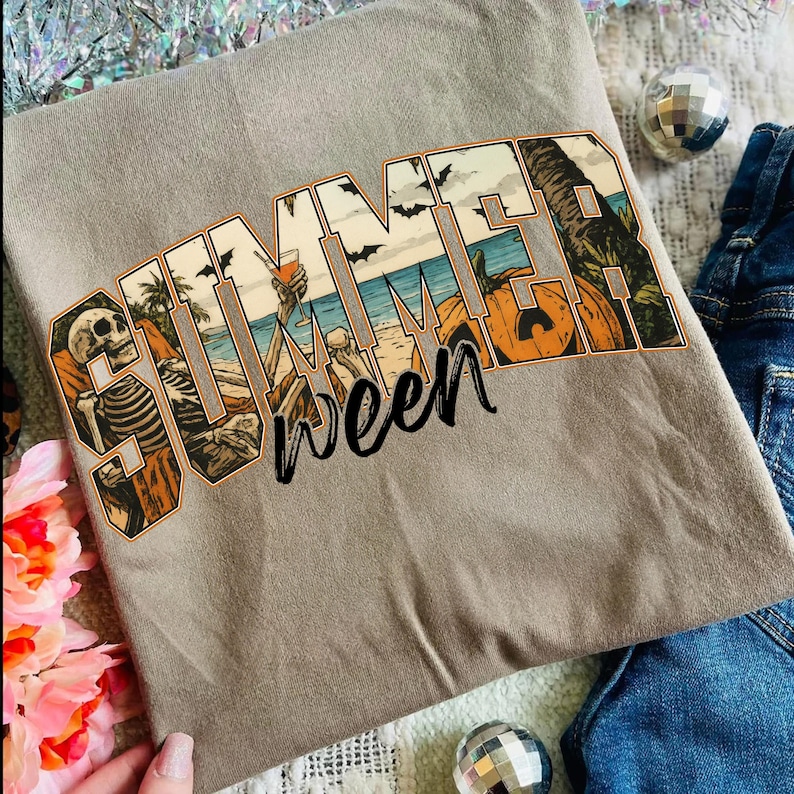 May include: A light brown t-shirt with the word "SUMMERWEEN" in a colorful graphic. The graphic features a skeleton, beach scene, palm trees, bats, and pumpkins. The shirt is folded and placed on a surface with decorative elements.