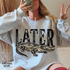 Peut inclure: Un sweat-shirt gris clair avec le mot "LATER" en grandes lettres noires au-dessus d'une illustration d&eacute;taill&eacute;e d'un alligator. Le sweat-shirt a un col rond et des manches longues. Le logo Infinite Pixel Studio est en bas &agrave; gauche.