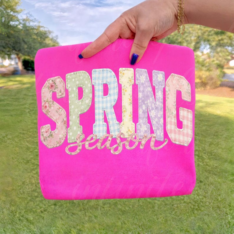 May include: A bright pink sweatshirt with the word "SPRING" in patchwork letters. The letters are in various floral and gingham patterns. The word "season" is embroidered below in a cursive font. The sweatshirt is held up by a hand.