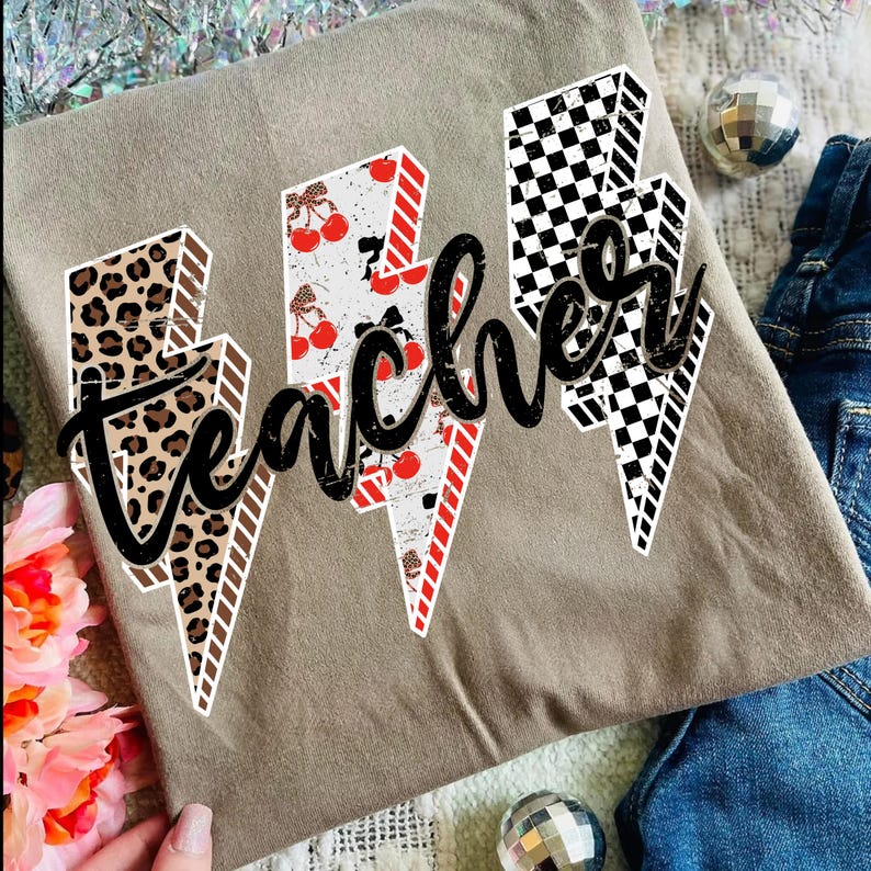 May include: A tan t-shirt featuring three lightning bolt designs with the word "teacher" in black script. The lightning bolts have different patterns: leopard print, cherries, and a black and white checkerboard.
