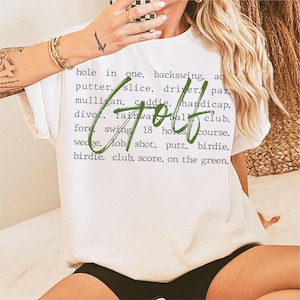 May include: White t-shirt with green text that says "Golf" and a list of golf terms including "hole in one", "backswing", "putter", "slice", "driver", "par", "mulligan", "caddie", "handicap", "divot", "fairway", "ball", "club", "fore", "swing", "18 holes", "course", "wedge", "lob shot", "putt", "birdie", "birdie", "club", "score", "on the green".