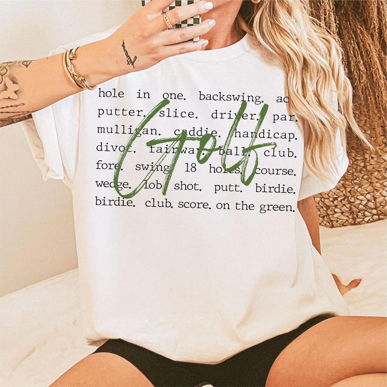 May include: White t-shirt with green text that says "Golf" and a list of golf terms including "hole in one", "backswing", "putter", "slice", "driver", "par", "mulligan", "caddie", "handicap", "divot", "fairway", "ball", "club", "fore", "swing", "18 holes", "course", "wedge", "lob shot", "putt", "birdie", "birdie", "club", "score", "on the green".