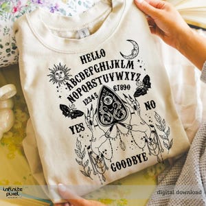 May include: Cream-coloured sweatshirt with a black Ouija board design. The design includes the alphabet, numbers, sun, moon, butterflies, and the words "Hello", "Yes", "No", and "Goodbye". The design features hands holding a planchette.