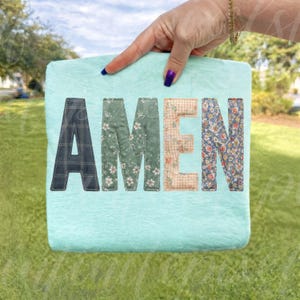 May include: A light blue square blanket with the word "AMEN" stitched across the front. The letters are made of various patterned fabrics, including plaid, floral, and gingham. The blanket is held up by a hand with a gold bracelet and purple nail polish.