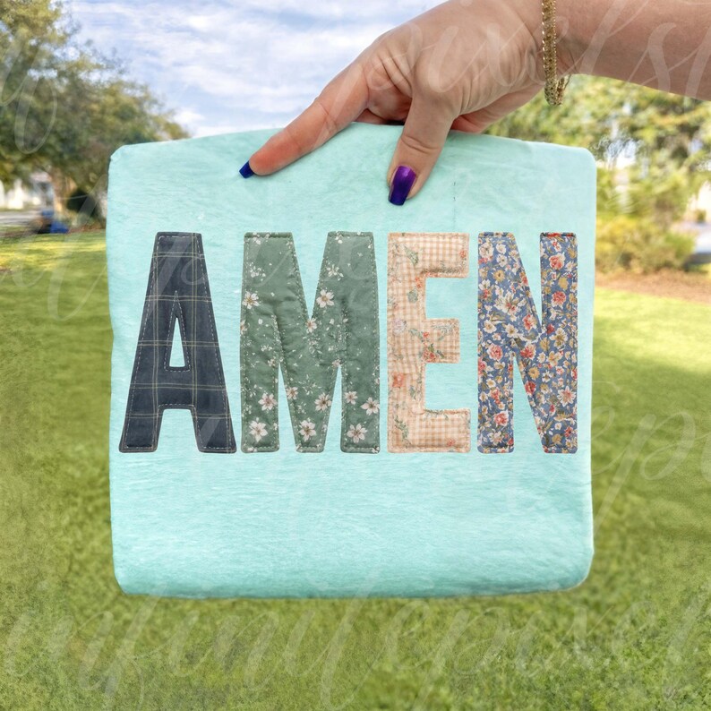 May include: A light blue square blanket with the word "AMEN" stitched across the front. The letters are made of various patterned fabrics, including plaid, floral, and gingham. The blanket is held up by a hand with a gold bracelet and purple nail polish.